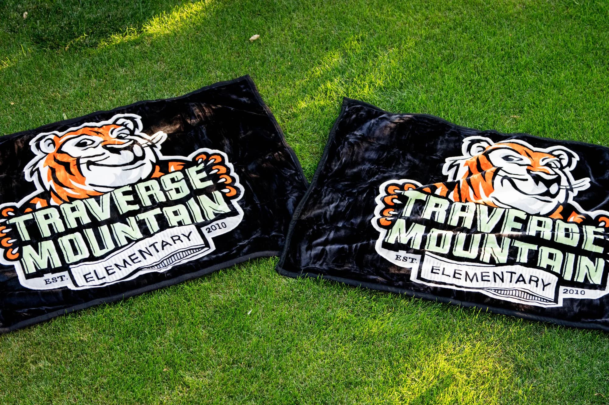 Two Traverse Mountain Elementary tiger mascot blankets laid out on grass, two layouts side by side