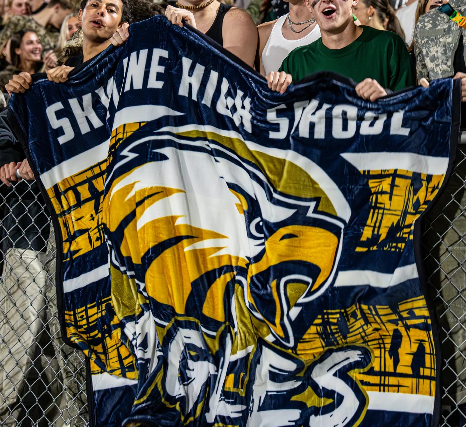 Students in the stands holding up a Skyline High School eagle mascot blanket at a night game